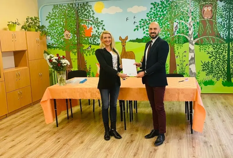 Zdjęcie o orientacji poziomej przedstawia dwie osoby stojące w sali przedszkolnej i trzymające wspólnie dokument w formacie A4. Kobieta po lewej stronie ubrana jest w czarną marynarkę, jeansy i ciemne botki, natomiast mężczyzna po prawej ma na sobie elegancką marynarkę, białą koszulę z krawatem oraz bordowe spodnie. Przed nimi znajduje się długi stół przykryty brzoskwiniowym obrusem, na którym stoi wazon z bukietem czerwono-białych kwiatów. Tło stanowi kolorowy mural z motywami leśnymi, przedstawiający drzewa, sarnę oraz sowę, co wskazuje na przyjazne środowisko edukacyjne. Całość kadru, utrzymana w ciepłej kolorystyce, doskonale nadaje się do ilustracji artykułów o współpracy partnerskiej, inwestycjach w placówki oświatowe lub oficjalnych podpisaniach umów.