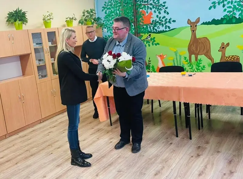 Zdjęcie przedstawia scenę gratulacji w pomieszczeniu o charakterze edukacyjnym lub przedszkolnym, utrzymaną w orientacji poziomej. W centrum kadru kobieta o jasnych włosach, ubrana w czarną marynarkę i ciemne jeansy, ściska dłoń mężczyzny w okularach i szarej marynarce, który trzyma bukiet białych chryzantem z czerwonymi różami. W tle widoczny jest inny mężczyzna w ciemnym swetrze oraz stół przykryty brzoskwiniowym obrusem, ustawiony na tle ściany ozdobionej malowidłem ściennym przedstawiającym las z jeleniami i lisem. Po lewej stronie znajdują się wysokie, jasne szafki biurowe z roślinami doniczkowymi na blatach. Podłoga wyłożona jest jasnym panelem o fakturze drewna, co nadaje wnętrzu ciepły, przyjazny klimat. Cała kompozycja jest dobrze doświetlona i ma charakter oficjalnego, lokalnego wydarzenia.