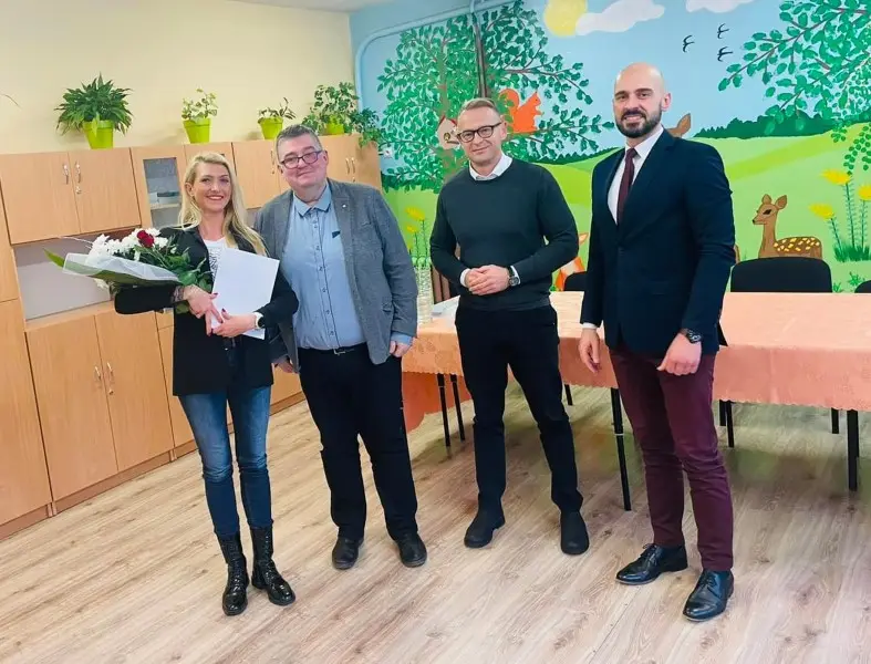 Zdjęcie wykonane w orientacji poziomej przedstawia cztery uśmiechnięte osoby stojące w sali przedszkolnej lub szkolnej. Po lewej stronie stoi blondynka w czarnym żakiecie i jeansach, trzymająca bukiet kwiatów oraz białą teczkę. Obok niej znajduje się trzech mężczyzn: jeden w szarej marynarce, drugi w ciemnozielonym swetrze i trzeci w eleganckim, granatowym garniturze z bordowym krawatem. Tło stanowi jasna ściana ozdobiona kolorowym malowidłem ściennym z motywami leśnych zwierząt, takimi jak sarna i wiewiórka. Po lewej stronie zdjęcia widać rząd drewnianych szafek z roślinami doniczkowymi na górze, a na pierwszym planie stoi stół przykryty łososiowym obrusem. Kompozycja zdjęcia jest symetryczna, a postacie stanowią centralny punkt kadru w jasnym, przytulnym wnętrzu.