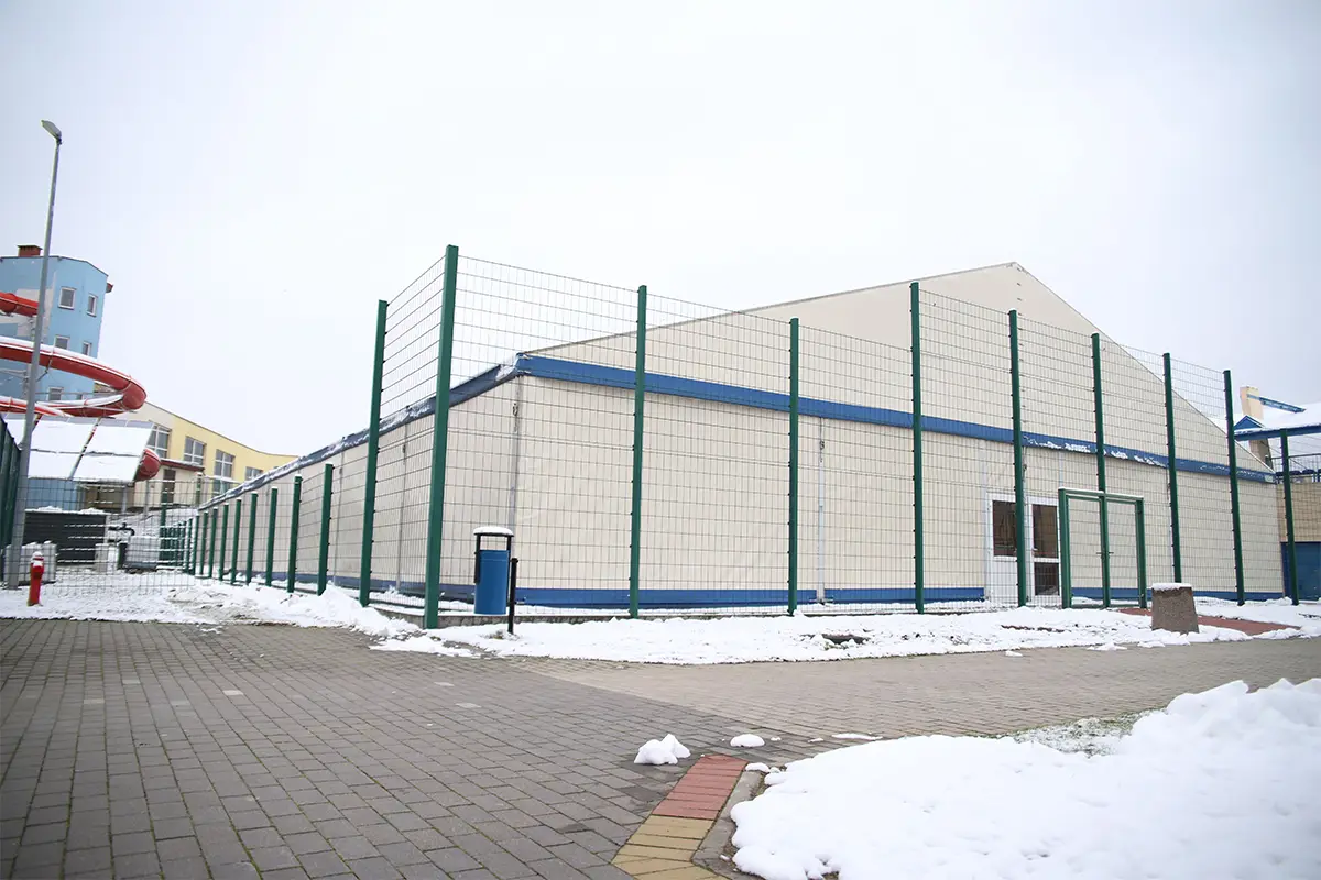 Zdjęcie wykonane w orientacji poziomej przedstawia dużą halę sportową o konstrukcji namiotowej, otoczoną wysokim metalowym ogrodzeniem panelowym w kolorze ciemnozielonym. Budynek posiada jasne ściany z wyraźnymi niebieskimi akcentami wzdłuż linii dachu oraz przy podstawie, a na pierwszym planie widoczny jest chodnik wyłożony kostką brukową pokrytą miejscami warstwą śniegu. Po lewej stronie w tle majaczy fragment niebieskiego budynku z czerwoną rurą zjeżdżalni, co sugeruje lokalizację w pobliżu kompleksu basenowego lub aquaparku. Niebo nad obiektem jest zachmurzone i jasne, co tworzy równomierne, rozproszone oświetlenie całej sceny. Całość kadru ma charakter dokumentalny, prezentując architekturę sportową w zimowej aurze.