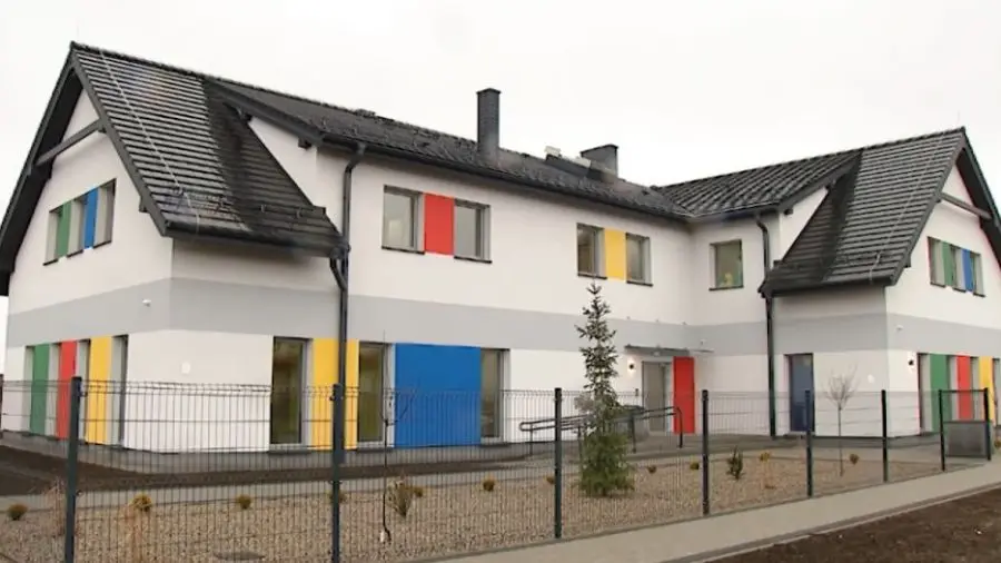Zdjęcie przedstawia nowoczesny budynek użyteczności publicznej lub przedszkole w układzie poziomym o proporcjach boków zbliżonych do 16:9. Architektura obiektu utrzymana jest w jasnej kolorystyce, którą przełamują żywe akcenty w kolorach niebieskim, czerwonym, żółtym i zielonym, umieszczone między oknami oraz przy wejściu. Budynek posiada dwuspadowy dach kryty ciemną dachówką oraz charakterystyczny poziomy szary pas przebiegający przez elewację. Całość terenu otoczona jest metalowym ogrodzeniem z siatki, przed którym znajduje się chodnik, a w ogrodzie posadzono młode drzewko iglaste. Konstrukcja bryły jest rozłożysta, z wyraźnym podziałem na dwa skrzydła, co sugeruje przemyślany projekt funkcjonalnego obiektu edukacyjnego.